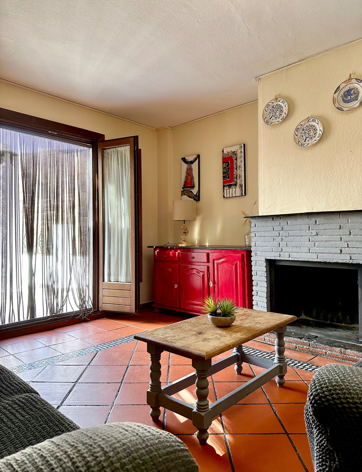 A cozy living area is presented with a brick fireplace and an inviting red cabinet. Natural light filters through sheer curtains, while a wooden table in the center holds a small potted plant. The tiled floor adds warmth to the space.