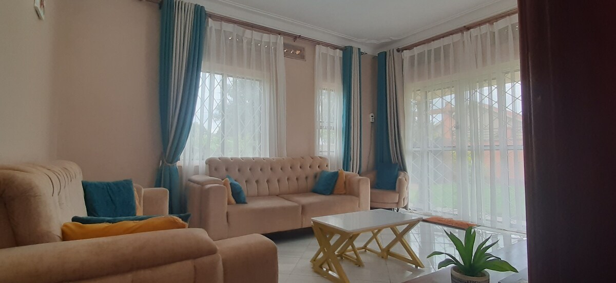 A light and airy living room is featured, with large windows draped in sheer curtains allowing natural light to enhance the space. Comfortable sofas in beige and teal are arranged around a modern yellow coffee table, complemented by a small green plant.