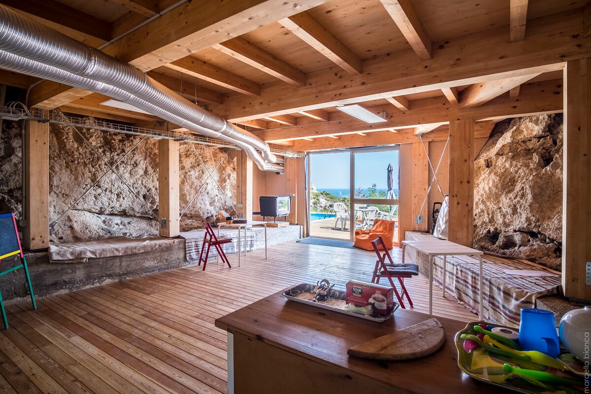 An open space features wooden beams and a polished wooden floor, accented by rock walls. Large glass doors lead to a view of the sea and a distant lighthouse. A small kitchen area is visible, alongside colorful chairs arranged for flexibility.