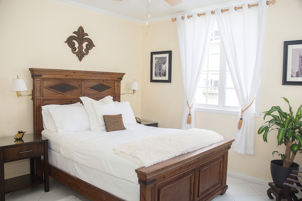 The private room features a wooden bed frame with a comfortable queen-sized bed, adorned with white linens and a decorative throw. Natural light enters through large windows, which are adorned with sheer white curtains. A bedside table and a plant add to the soothing ambiance.