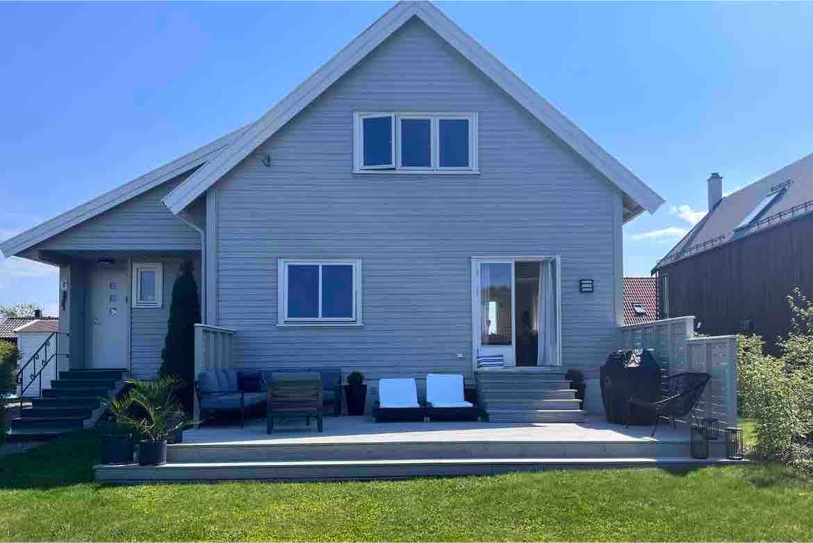 A light-colored wooden house features large windows and a spacious deck. Comfortable seating options are arranged on the deck, with a small table and greenery accentuating the outdoor space. The structure is positioned among a well-maintained lawn, inviting relaxation.