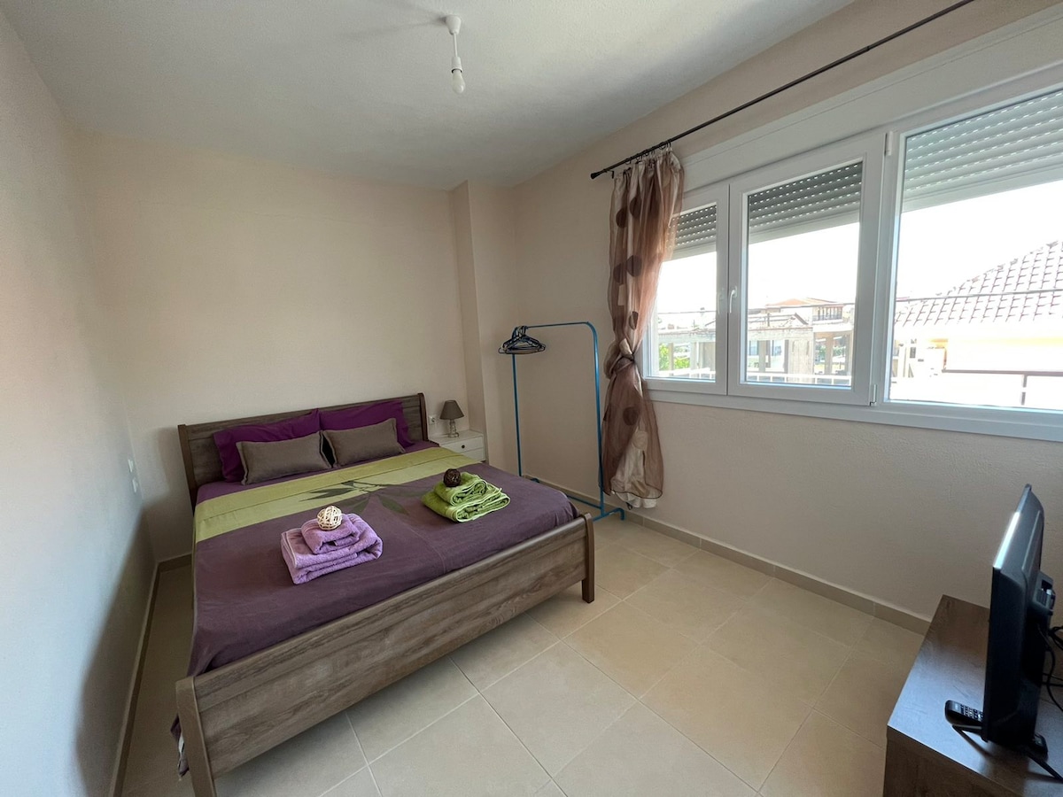 The bedroom features a spacious bed with a layered arrangement of sheets and towels in complementary colors. Natural light enters through large windows adorned with sheer curtains. A television sits on a wooden bedside unit, while a clothing rack provides additional storage.