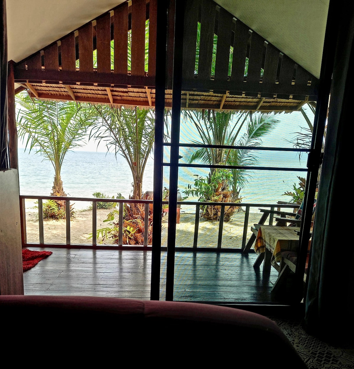 A serene view is captured through glass doors, showcasing palm trees on a sandy beach. The gentle waves can be seen in the distance, with vibrant greenery framing the scene, creating a connection between the indoors and the natural surroundings.