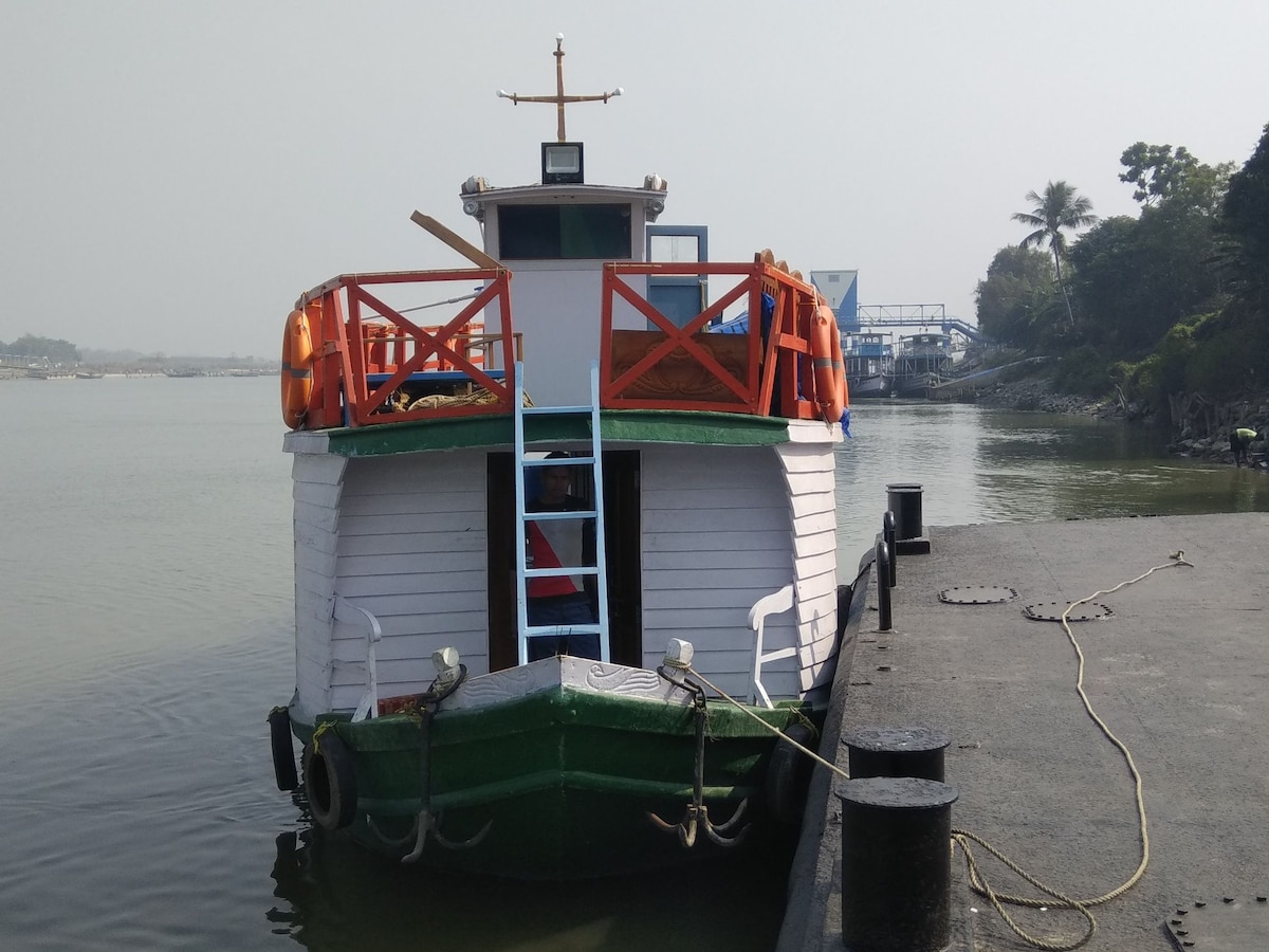 A colorful Eco-friendly houseboat with bright red railings and green trim is docked along a tranquil riverside. The boat features an accessible staircase leading to the upper deck, where stunning views of the water and surrounding nature can be enjoyed.
