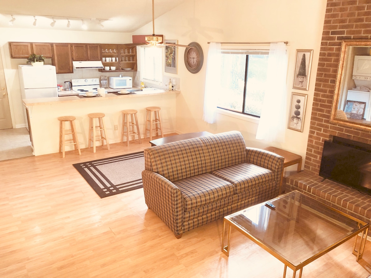 The open living area showcases a plaid sofa paired with a glass coffee table. A warm-toned wooden floor is complemented by a large area rug. The kitchen area is visible in the background, featuring light cabinetry and bar stools along the counter.