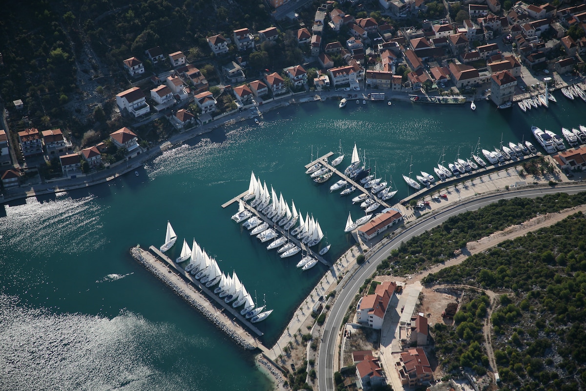 An aerial view captures a marina surrounded by a charming town, with numerous sailboats docked in the water. The harbor is lined with white sails, and terracotta-roofed buildings are nestled against a backdrop of green hills.