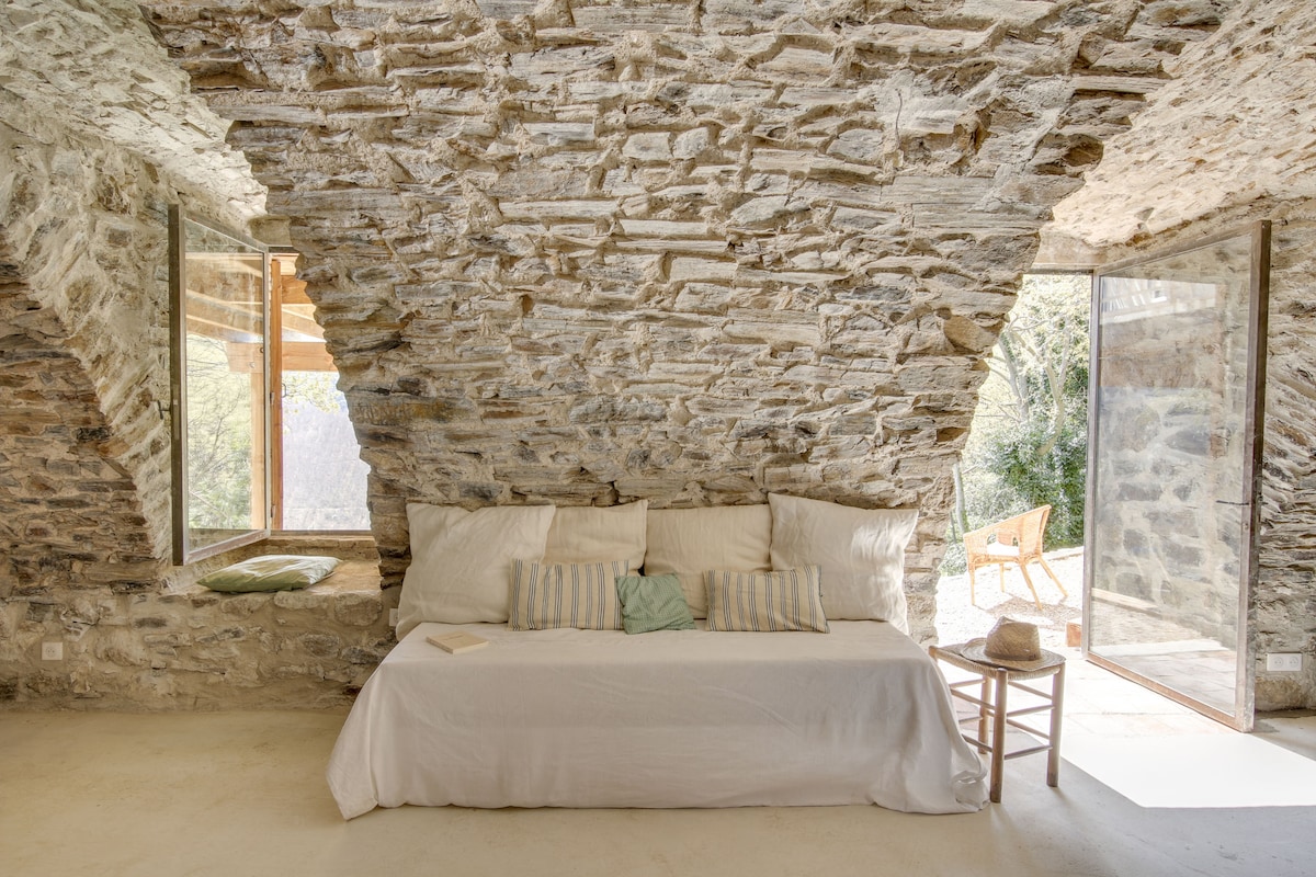 A spacious living area is highlighted by a white sofa adorned with various cushions. Natural light filters through large windows, complementing the rustic stone walls and ceiling. One window opens to the outdoors, offering a view of the surrounding greenery.