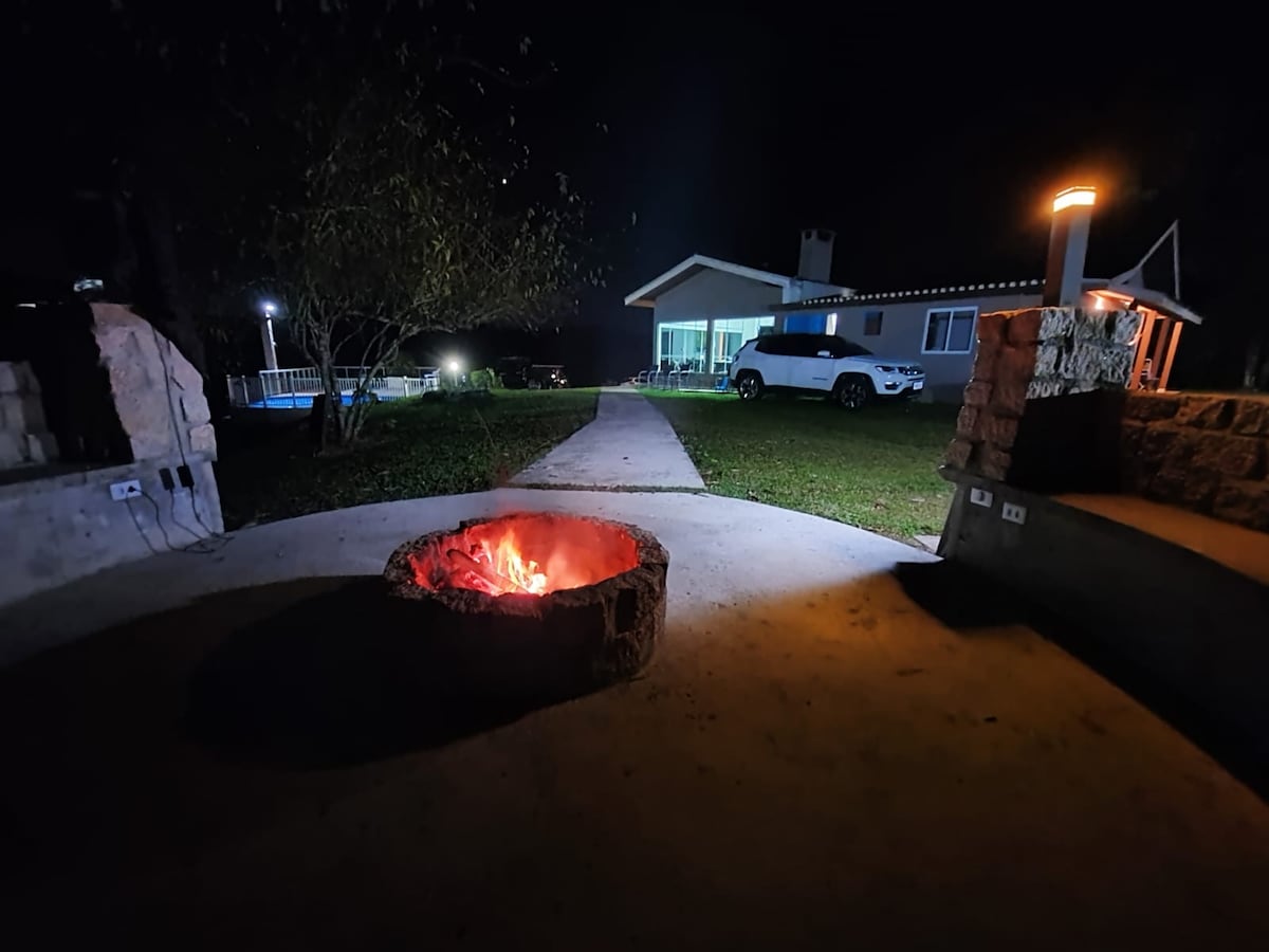 A stone fire pit with glowing embers is positioned on a concrete pathway. In the background, a well-lit house is visible among the trees, alongside a parked vehicle. Soft outdoor lighting creates a warm ambiance throughout the space.