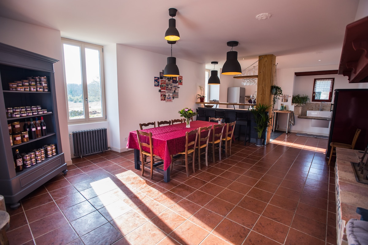 A spacious dining area features a long table set with a red tablecloth and surrounded by wooden chairs. Natural light streams through large windows, illuminating the room, while a well-equipped kitchen is visible in the background. Pantry shelves filled with jars are positioned against the wall.