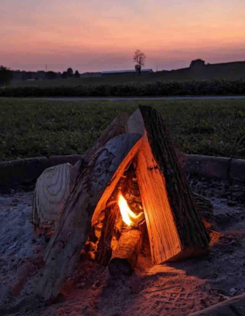 A campfire is set within a circular stone ring, with logs arranged in a tepee formation. A warm flame glows brightly in the center, surrounded by a sandy base. The sky above transitions from soft pinks to deep blues at dusk, complementing the serene country landscape.