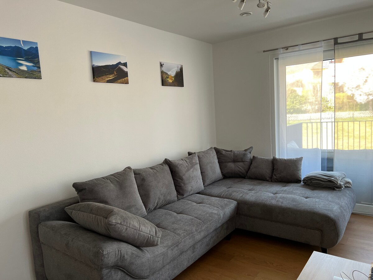 A spacious gray sectional sofa occupies one corner of the living area, offering ample seating. Above, three landscape photographs are displayed on the wall. Large windows allow natural light to fill the room, with a light curtain partially covering the view of the balcony.