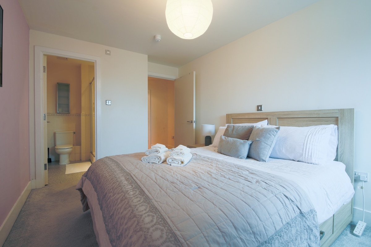 A spacious, well-lit main bedroom features a king-sized bed with luxurious white linens. Decorative pillows enhance the bedding. A wardrobe and chest of drawers offer storage, while light flows in from a nearby window. A cozy bathroom is visible through an open door.