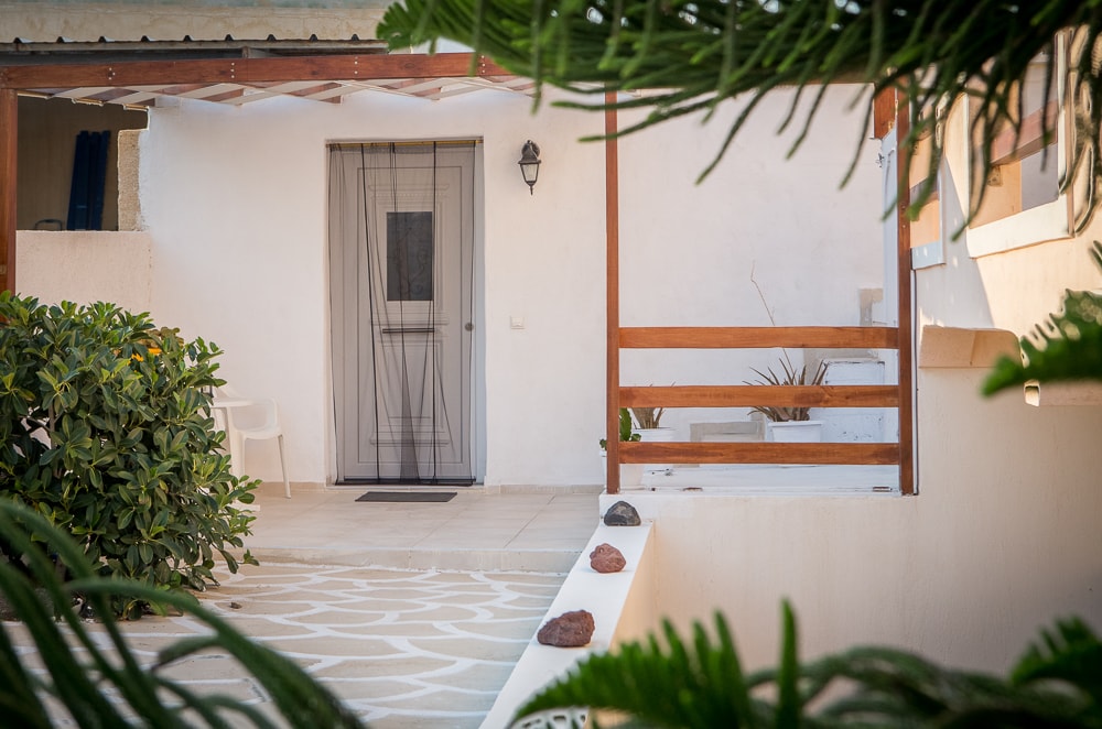 The entrance to the private guesthouse is framed by green foliage. A simple wooden structure with diagonal slats provides partial enclosure. The entrance features a neutral-colored door with a window and a welcome mat. A white chair is positioned nearby, along with natural stone accents along the pathway.