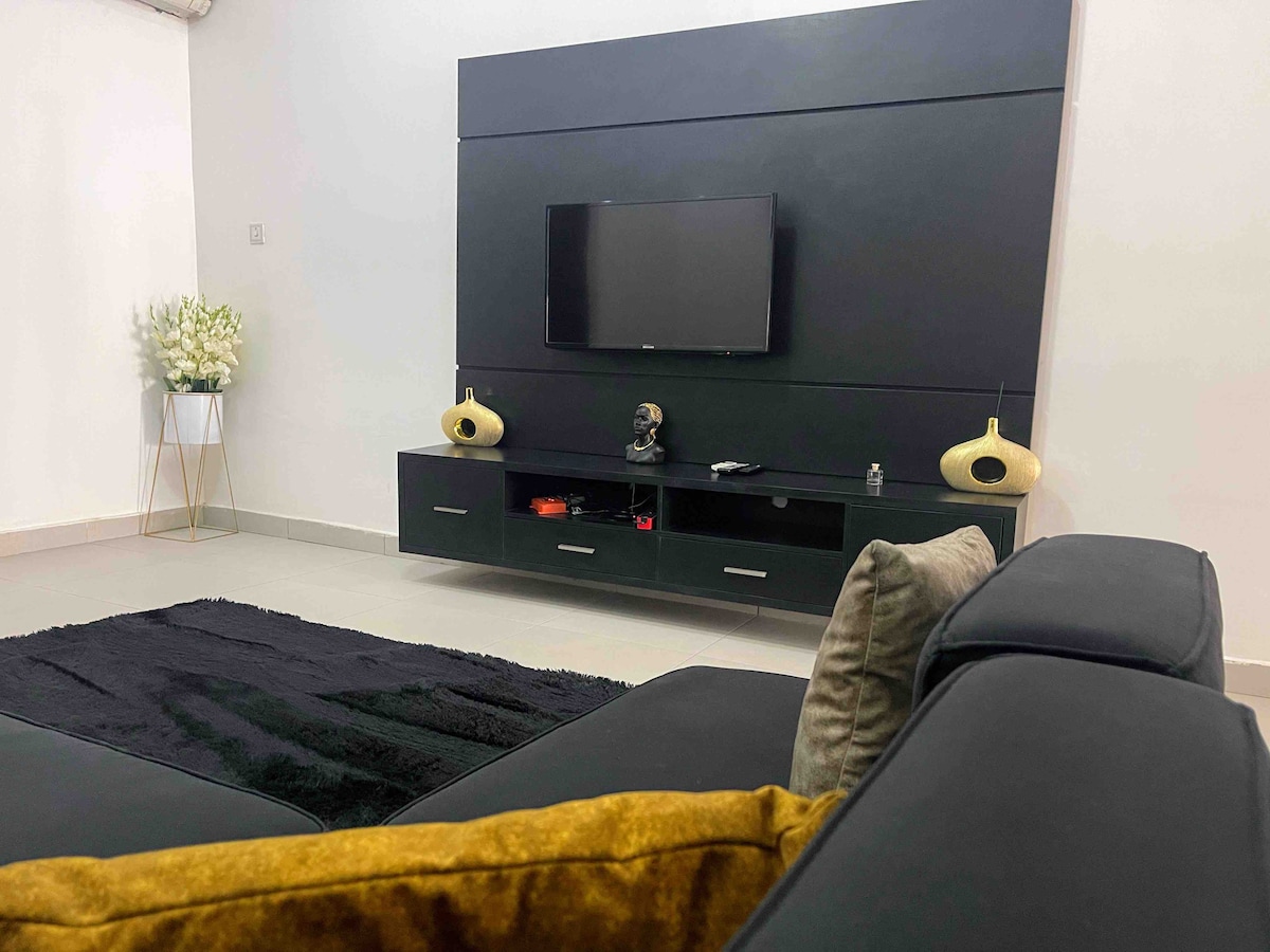 A modern living area showcases a sleek black entertainment unit featuring a flat-screen TV. Below, storage cabinets are paired with stylish decorative accents. A plush black rug lies underneath, while a corner table holds a small bouquet of flowers, contributing to a clean and minimal aesthetic.