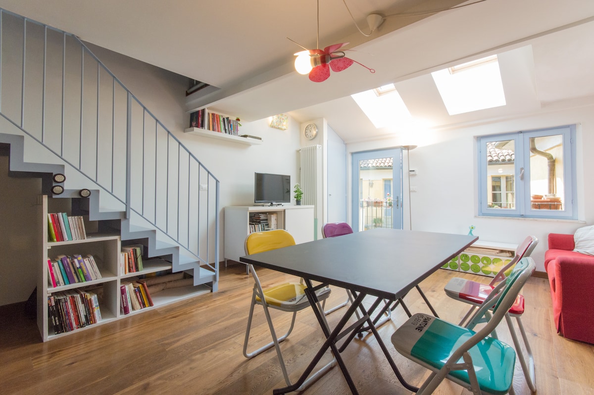 A cozy living area features a large table surrounded by colorful chairs. A small television is positioned on a white cabinet, while a bookshelf is filled with various books. Natural light pours in through two skylights and two windows, enhancing the airy atmosphere.