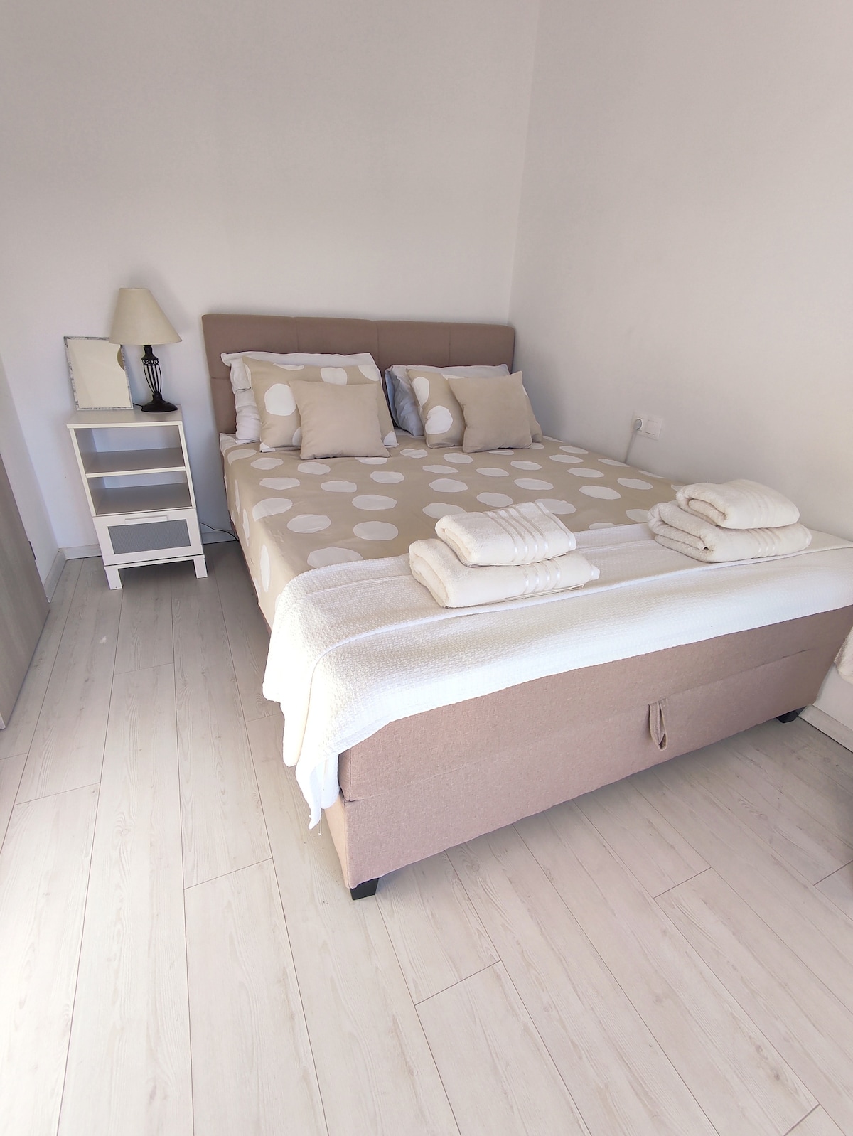 A comfortable bedroom features a spacious bed covered with a neutral polka dot blanket and flanked by two pillows. A bedside lamp and a nightstand with a drawer are visible, along with neatly arranged towels at the foot of the bed. Light-colored flooring adds to the bright atmosphere.