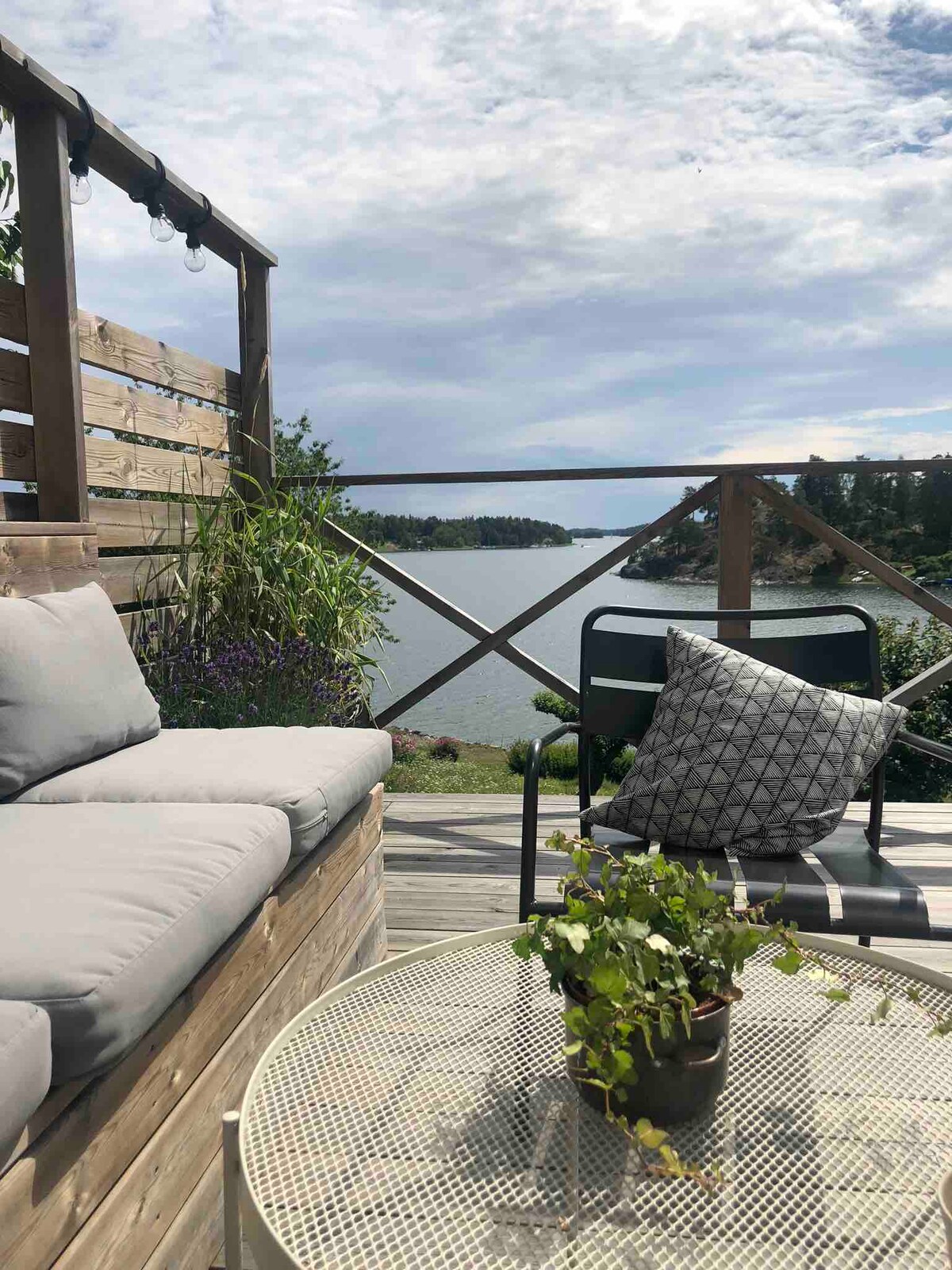 A balcony area is shown with comfortable seating, including a cushioned bench and a black chair. A small table holds a potted plant. The view overlooks the water and surrounding greenery, with clouds scattered across the sky.