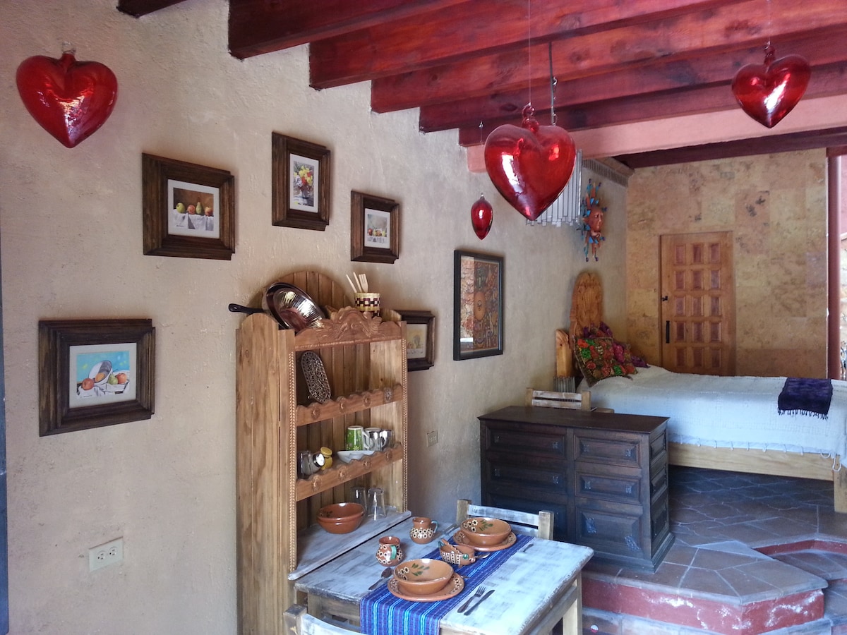 A rustic interior features wooden beams overhead and vibrant heart-shaped decorations hanging from the ceiling. A wooden dining table is set with traditional dishware, while framed artwork adorns the walls. A comfortable sleeping area with colorful textiles is visible in the background.