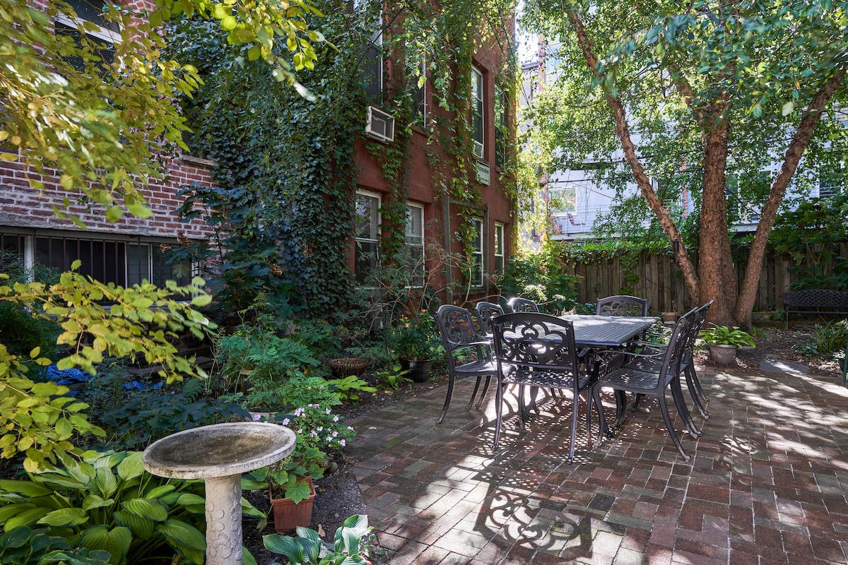 A tranquil garden area is surrounded by lush greenery and brick walls, featuring a stone patio with a table and six chairs. Mature trees provide shade, while various plants and flowers enhance the serene outdoor setting.