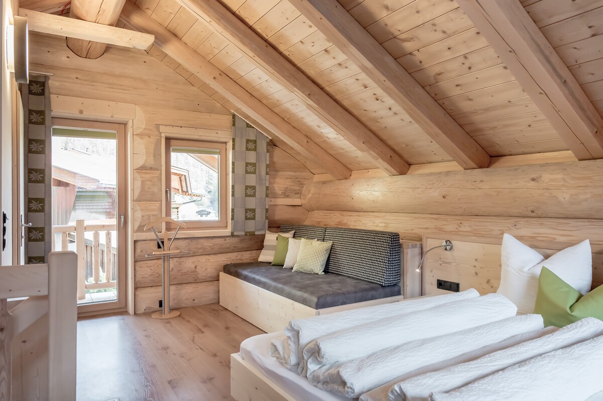 The interior of a chalet features wooden walls and a sloped ceiling with exposed beams. A spacious bed is positioned beside a large window, allowing natural light to enter. A cozy seating area with decorative cushions is visible, enhancing the comfort of the space.