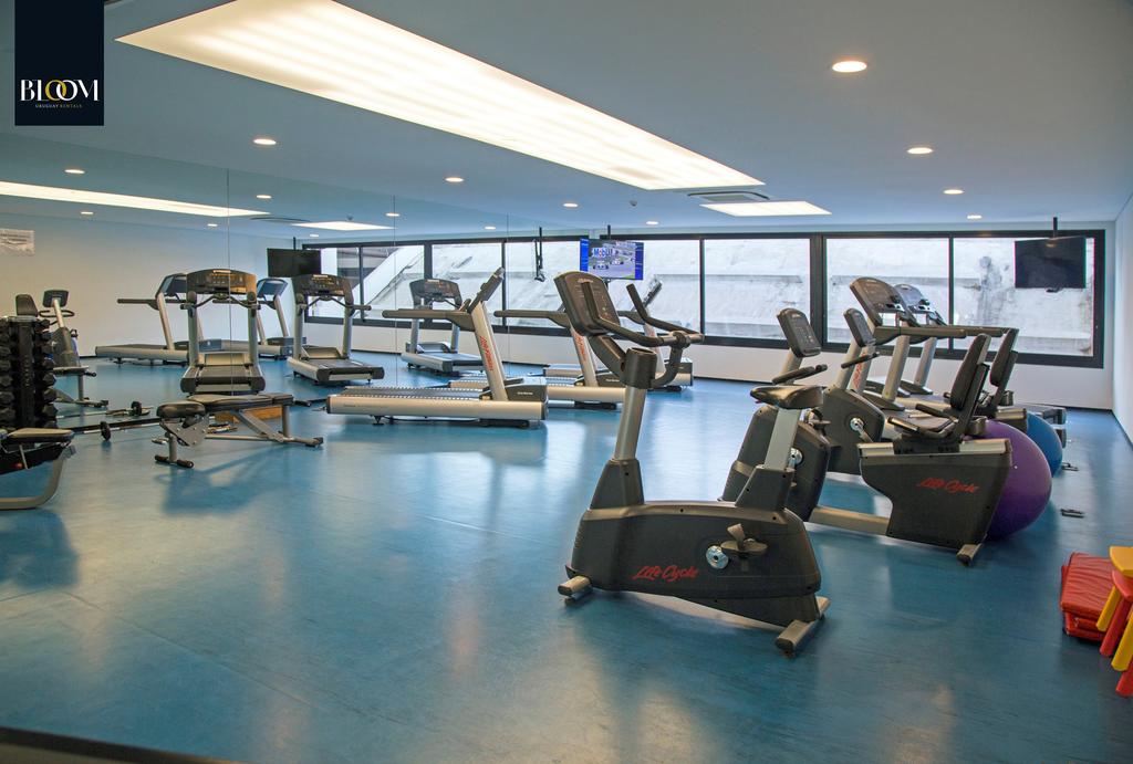 A well-equipped gym space is illuminated by natural and artificial lighting. Various exercise machines, including treadmills and stationary bikes, are arranged across a blue rubber floor. Large windows provide ample natural light, enhancing the overall exercise environment.