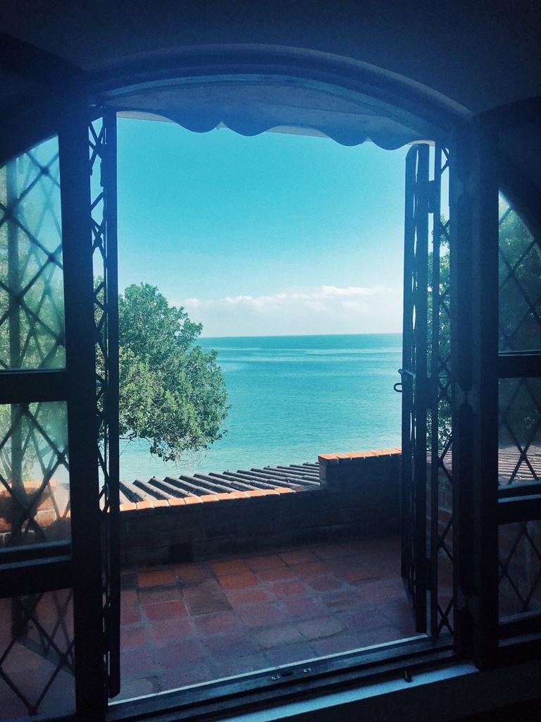 Arched windows frame a serene view of the Indian Ocean, with gentle waves lapping against the shore. The vibrant blue sky complements the tranquil water, while verdant trees add a touch of greenery to the scene, inviting guests to enjoy the coastal landscape.