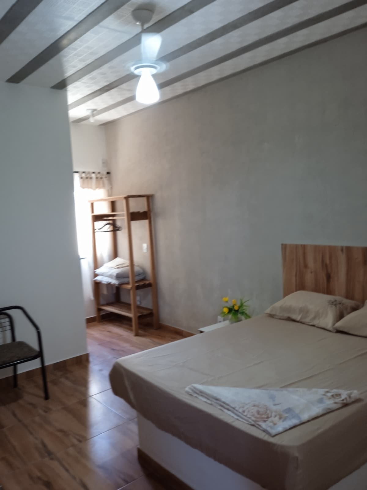 A spacious room is displayed, featuring a large bed with neutral-colored linens. A small arrangement of yellow flowers sits on the bedside. A chair is positioned in the corner, and an open wardrobe is seen in the background, providing ample storage space.