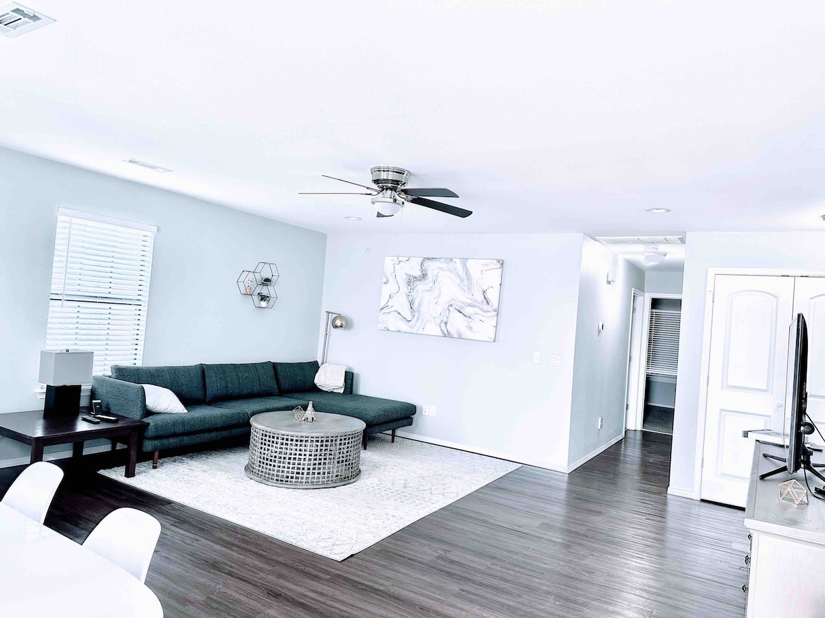 A spacious living room features a comfortable sectional sofa and a round coffee table at its center. A ceiling fan hangs overhead, and natural light enters through a window with blinds. A television is positioned against the wall, enhancing the entertainment space.
