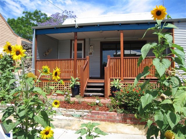 The front porch of the house is highlighted by sturdy wooden decking, featuring steps leading up to the entrance. Bright sunflowers and lush greenery frame the pathway, creating a welcoming atmosphere. The house has a blue trim, enhancing its charming appearance.