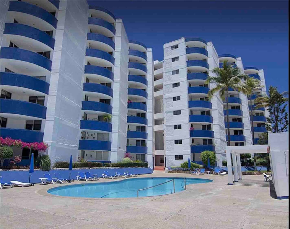 The image depicts a vibrant beachfront condo complex featuring multiple white and blue buildings. A clear pool is situated in the foreground, surrounded by lounge chairs and palm trees, with well-maintained gardens enhancing the inviting space.