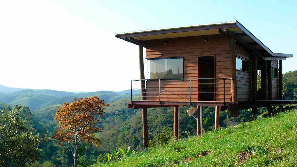 A wooden cabin is perched on stilts, offering expansive views of rolling green hills and a tree-lined valley. Large windows provide ample natural light, while a balcony extends from the structure, enhancing the connection to the surrounding nature.