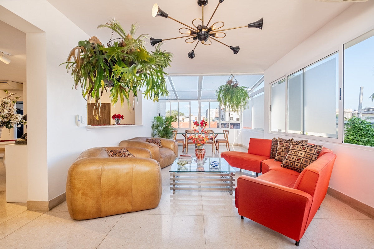 A bright and airy living area features a combination of brown and orange seating arrangements. Large windows allow natural light to illuminate the space, accented by potted plants hanging from the ceiling. A glass coffee table sits centrally, surrounded by decorative flowers.
