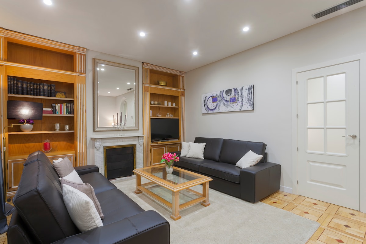 A spacious living area features two black leather sofas adjacent to a wooden coffee table, complemented by a decorative fireplace. A large mirror reflects light, while built-in shelving displays books and decorative items. A flat-screen TV is positioned for viewing pleasure.