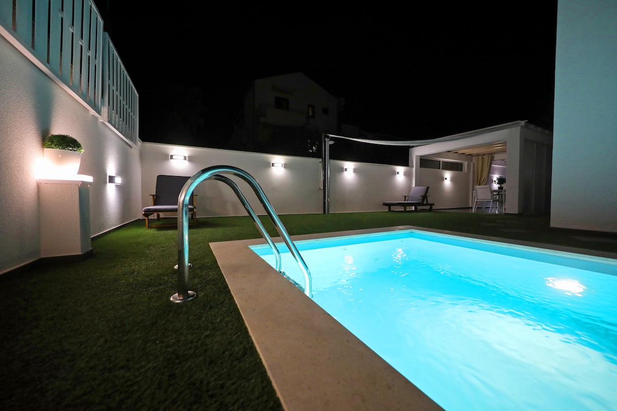 A heated outdoor swimming pool is illuminated softly, revealing crystal-clear water. Surrounding space features artificial grass, with lounge chairs positioned nearby. Soft lighting accents the white walls, creating a tranquil ambiance suited for evening relaxation.