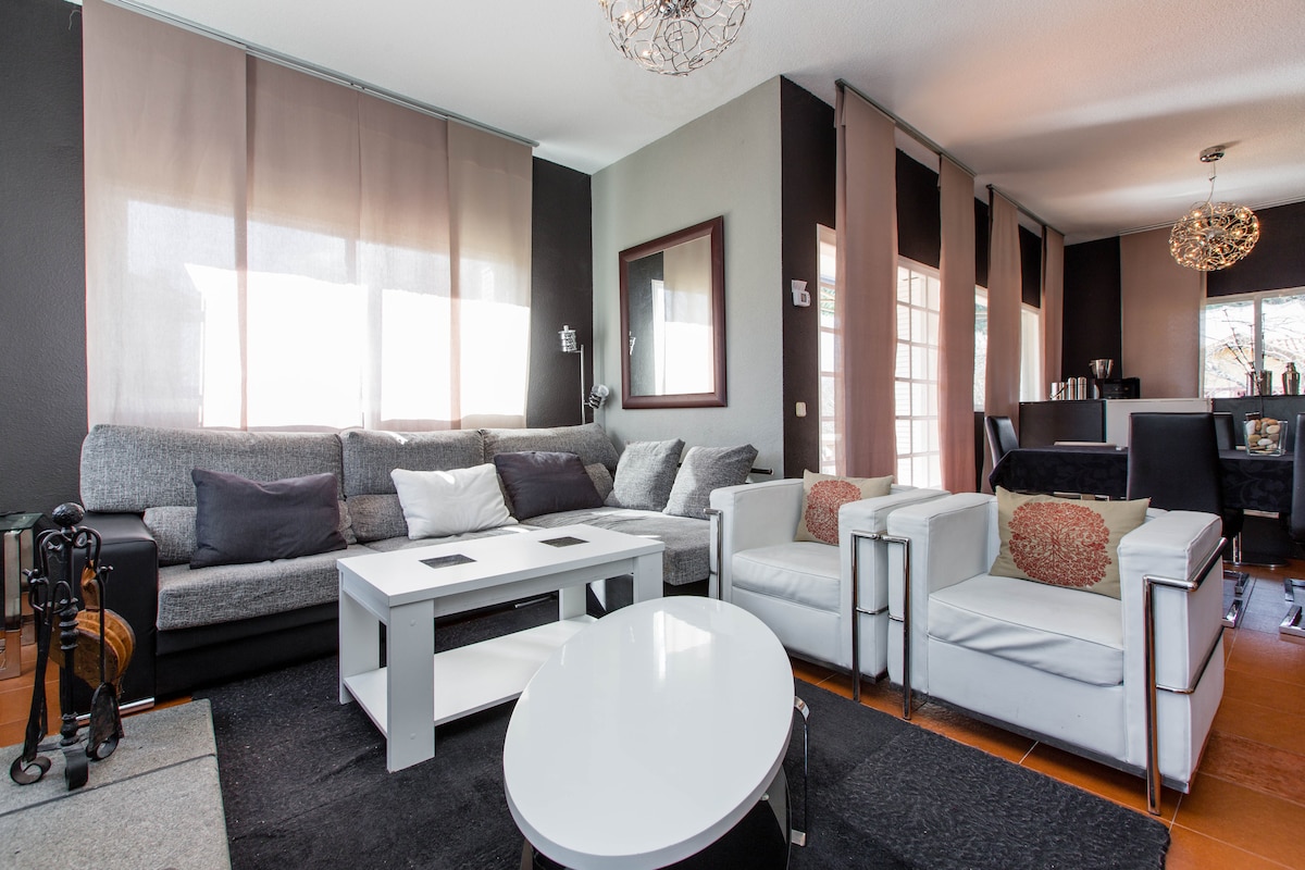 A spacious living area features a large gray sofa and white armchairs. Natural light enters through the large windows, accented by sheer curtains. A modern coffee table sits at the center, with a sleek dining area visible in the background.