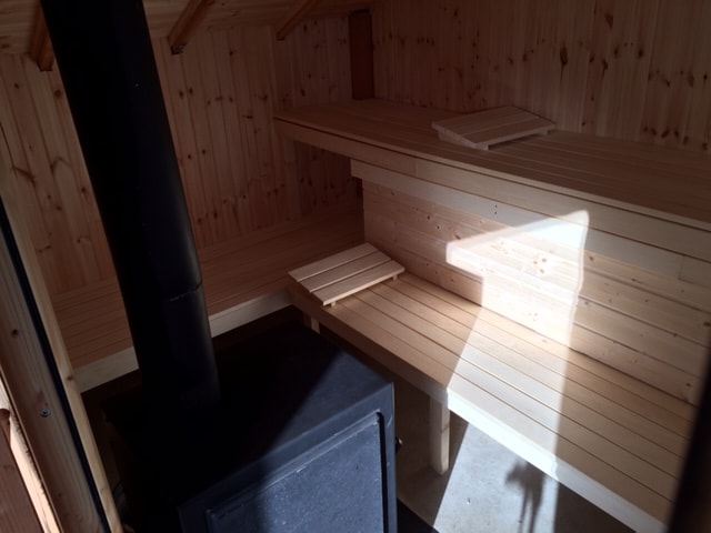 The interior of the wood-fired sauna features wooden benches arranged along the walls. Warm sunlight filters through a window, illuminating the natural wood tones. A black stove stands prominently on the side, enhancing the cozy atmosphere of the space.