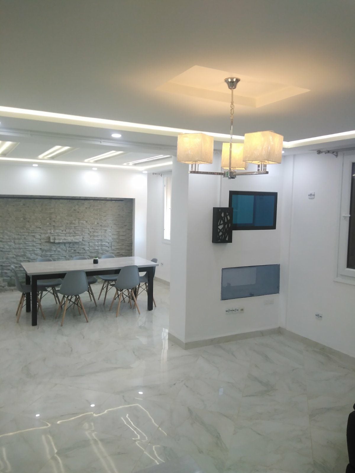 A contemporary dining area is presented, featuring a sleek table with a minimalist design surrounded by stylish chairs. A stone accent wall adds texture, while soft overhead lighting creates a warm ambiance in the bright space. Large windows allow for ample natural light.