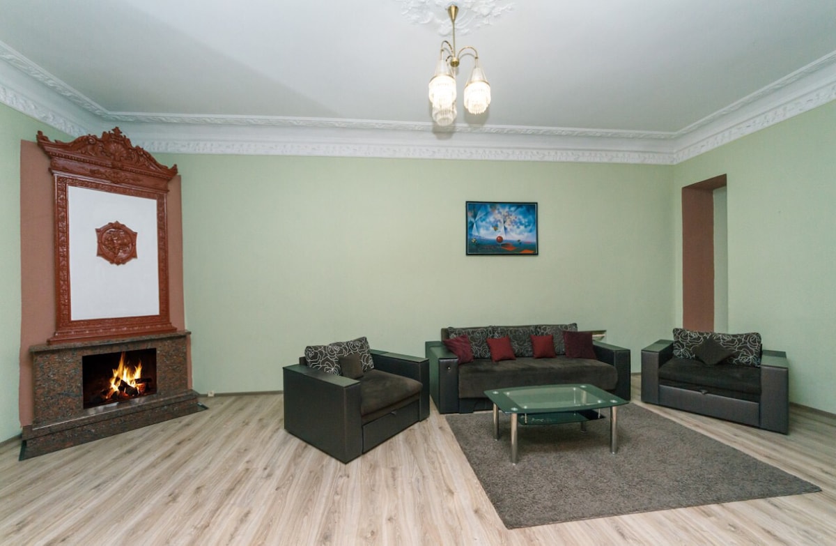 A spacious living area features a decorative fireplace set within an ornate mantel. Comfortable seating arrangements include a sofa and armchairs adorned with patterned cushions. A glass coffee table rests on a soft area rug, while light fixtures provide illumination in the room.