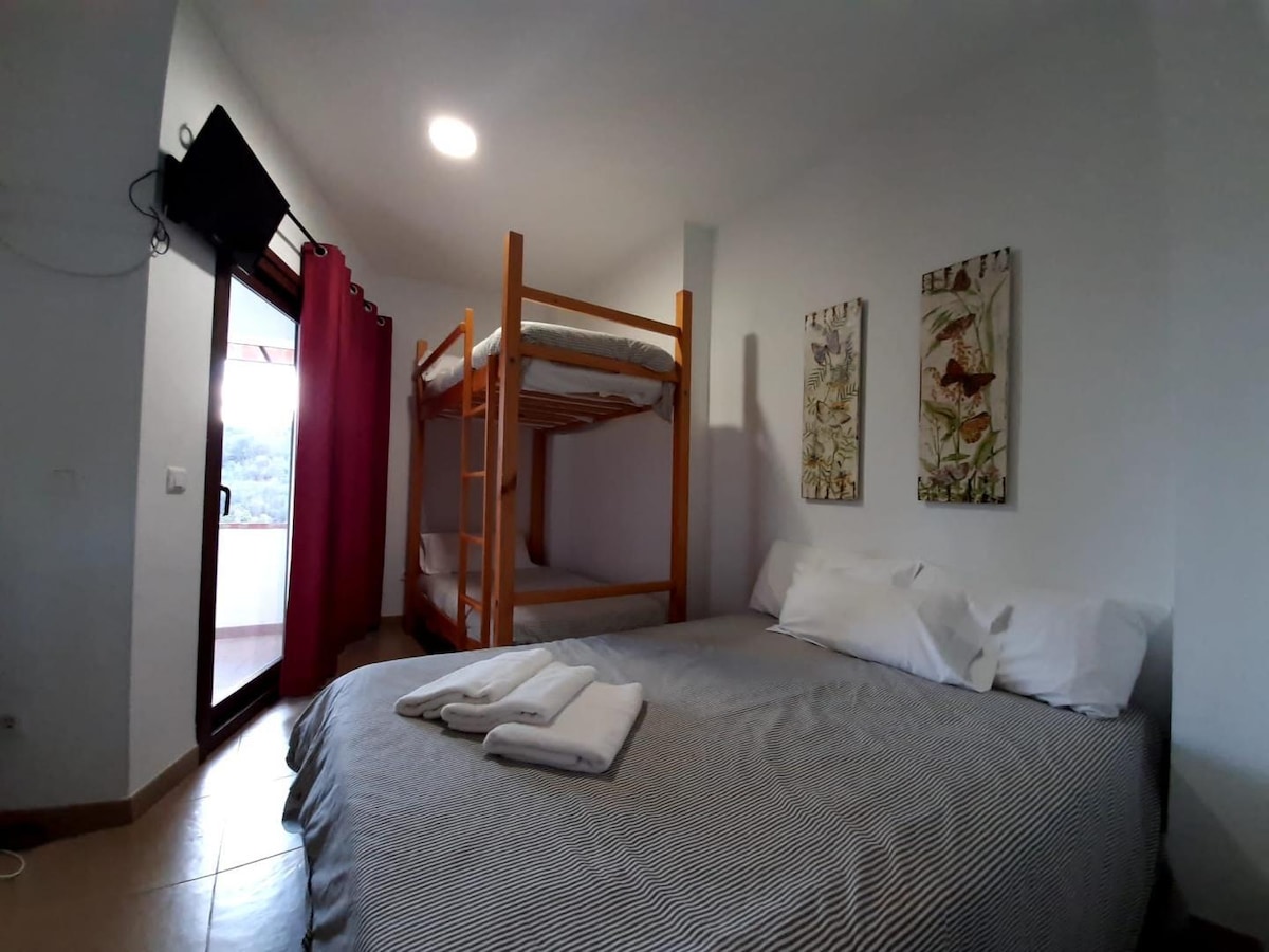A family room is presented with a spacious bed and a wooden bunk bed against the wall. Two floral art pieces are displayed above the bunk. Natural light enters through a glass door leading to a balcony, with a television mounted in the corner.