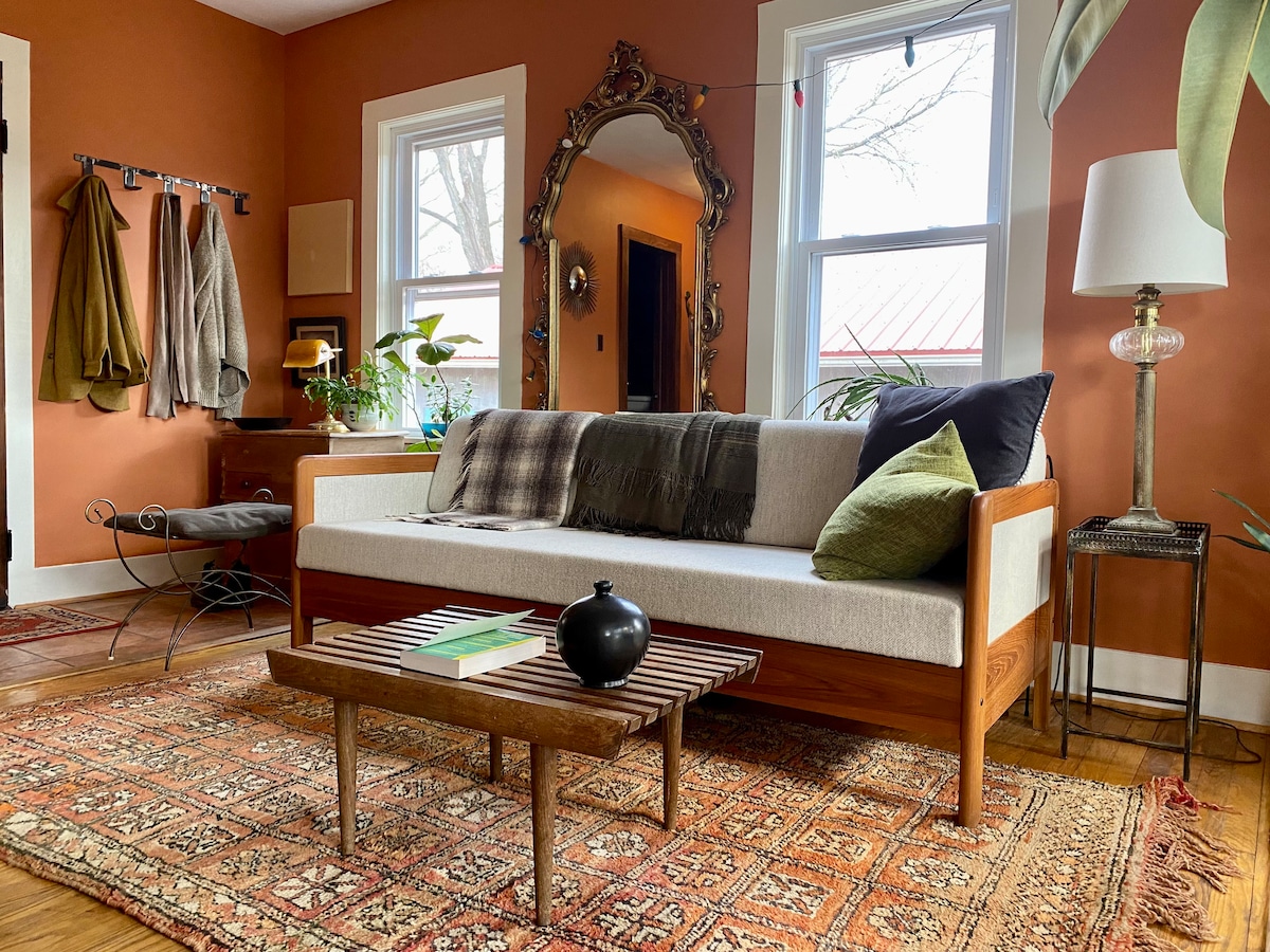 A living area features warm orange walls and a stylish sofa adorned with pillows. A low wooden table rests on a patterned rug, while plants provide a touch of greenery. Natural light streams in through windows, complemented by a vintage mirror and standing lamp.