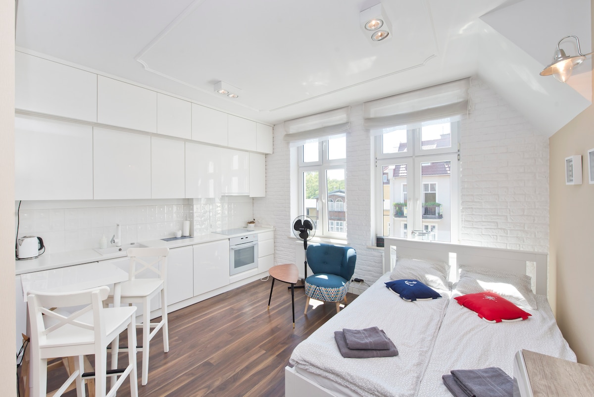 A modern studio apartment features a bright and open layout with large windows allowing natural light to fill the space. A spacious bed is positioned alongside a cozy seating area with a blue chair. A compact kitchen with white cabinetry offers functionality for guests.
