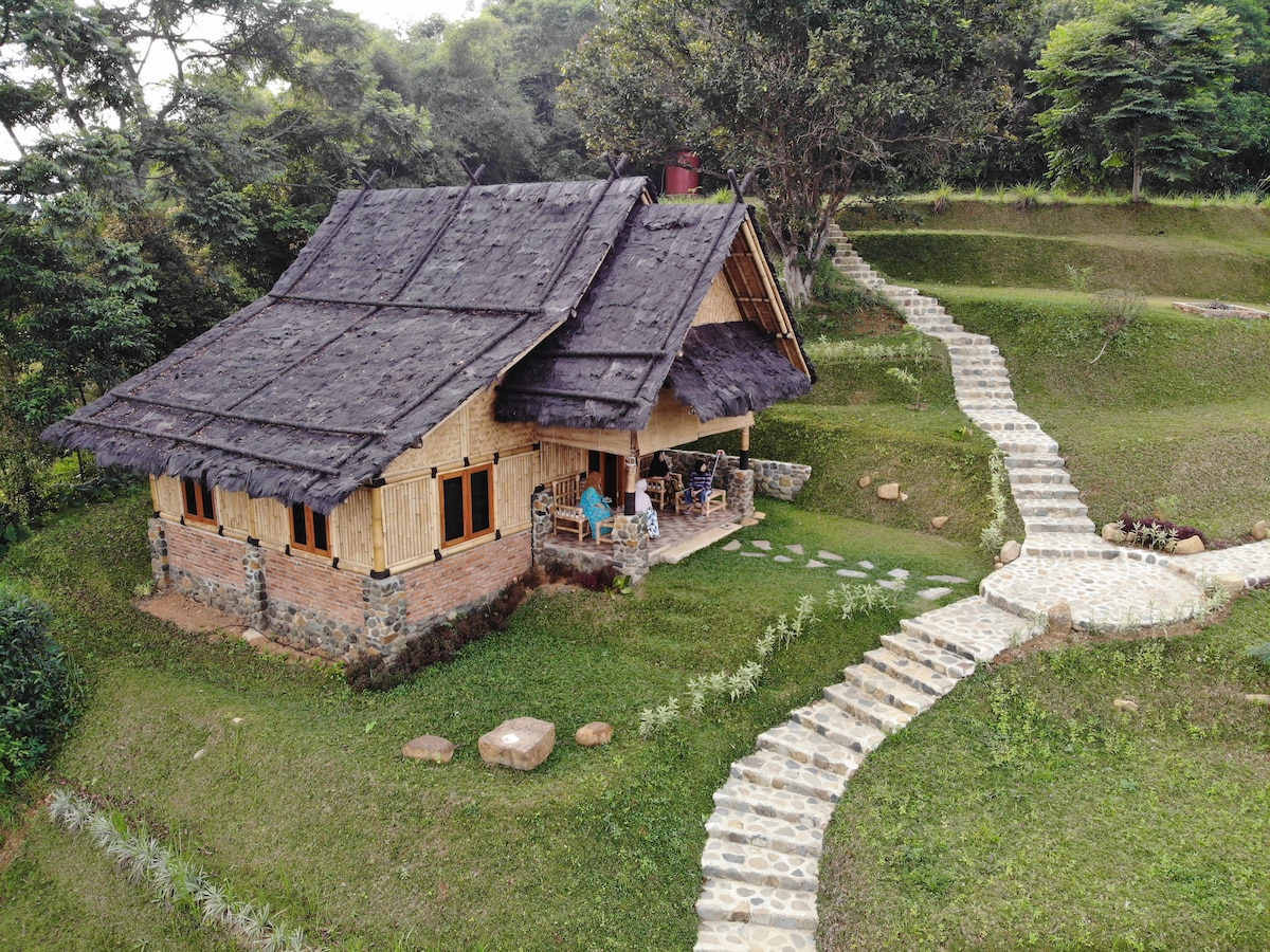 A bamboo cabin is nestled in a lush green landscape, featuring a thatched roof and stone accents. A pathway of stepping stones leads to the entrance, surrounded by a well-manicured lawn and small rocks, while trees provide a natural backdrop.
