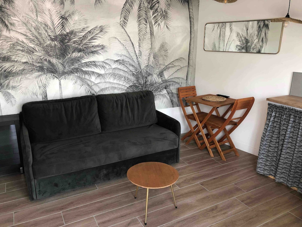 A modern living area is featured with a dark sofa facing a small round coffee table. Behind, a mural of palm trees adds a tropical touch. A compact dining set with wooden folding chairs is positioned next to a mirrored wall, enhancing the space's openness.