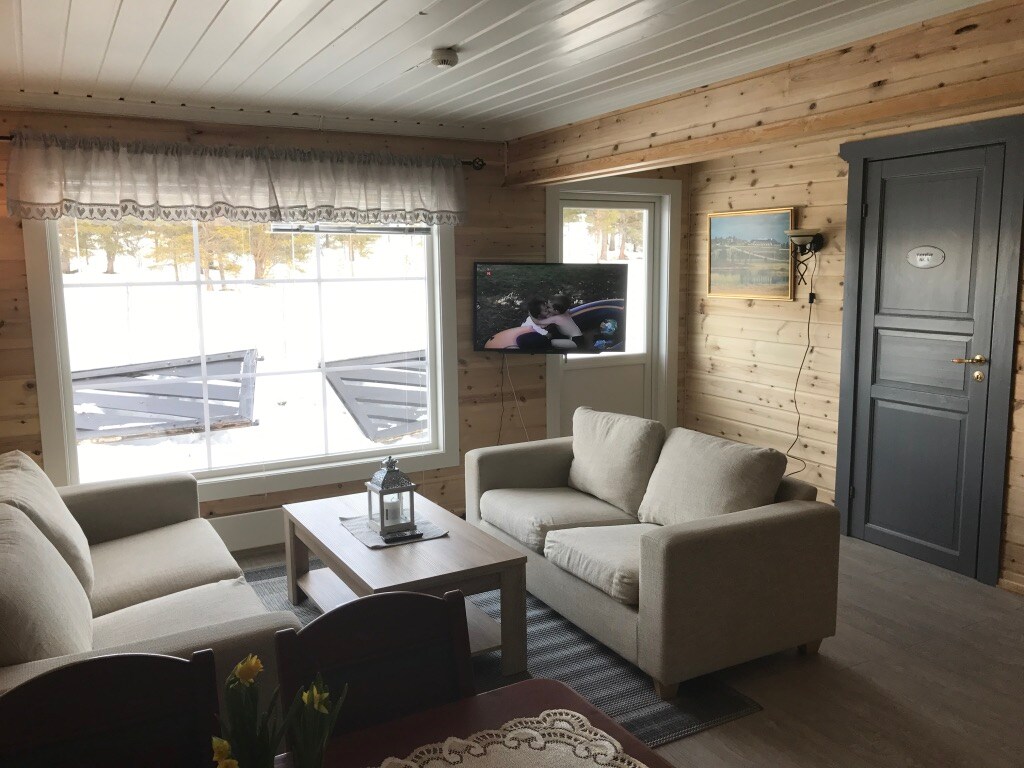 A cozy living area is shown, featuring two light-colored sofas arranged around a wooden coffee table. A television is mounted on the wall, and large windows allow natural light to fill the space, offering views of the outside snow-covered landscape.