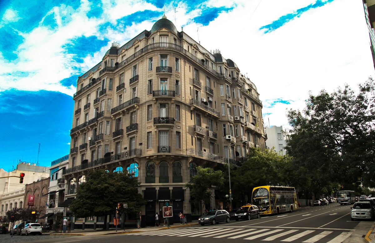 The historic building features intricate architectural details, with an elegant facade and a charming dome at the top. The structure is situated at a busy intersection, surrounded by trees and city life, showcasing the vibrant atmosphere of Buenos Aires.