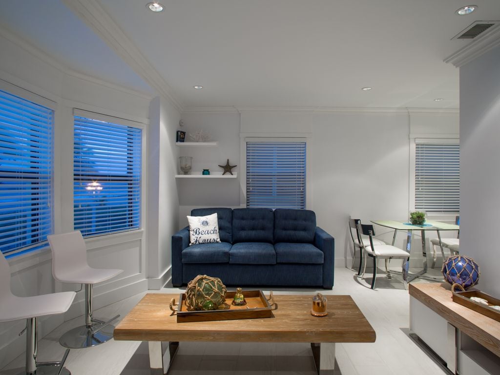 A modern living area features a navy blue sofa facing a wooden coffee table, surrounded by natural light from multiple windows. A glass dining table with metal chairs is positioned nearby, enhancing the open feel of the space.