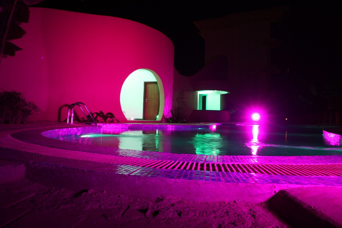 A modern swimming pool is illuminated with pink lighting at night, creating a vibrant atmosphere. The pool features a circular design with steps leading gently into the water. A doorway is visible in the background, hinting at the entry to the building.