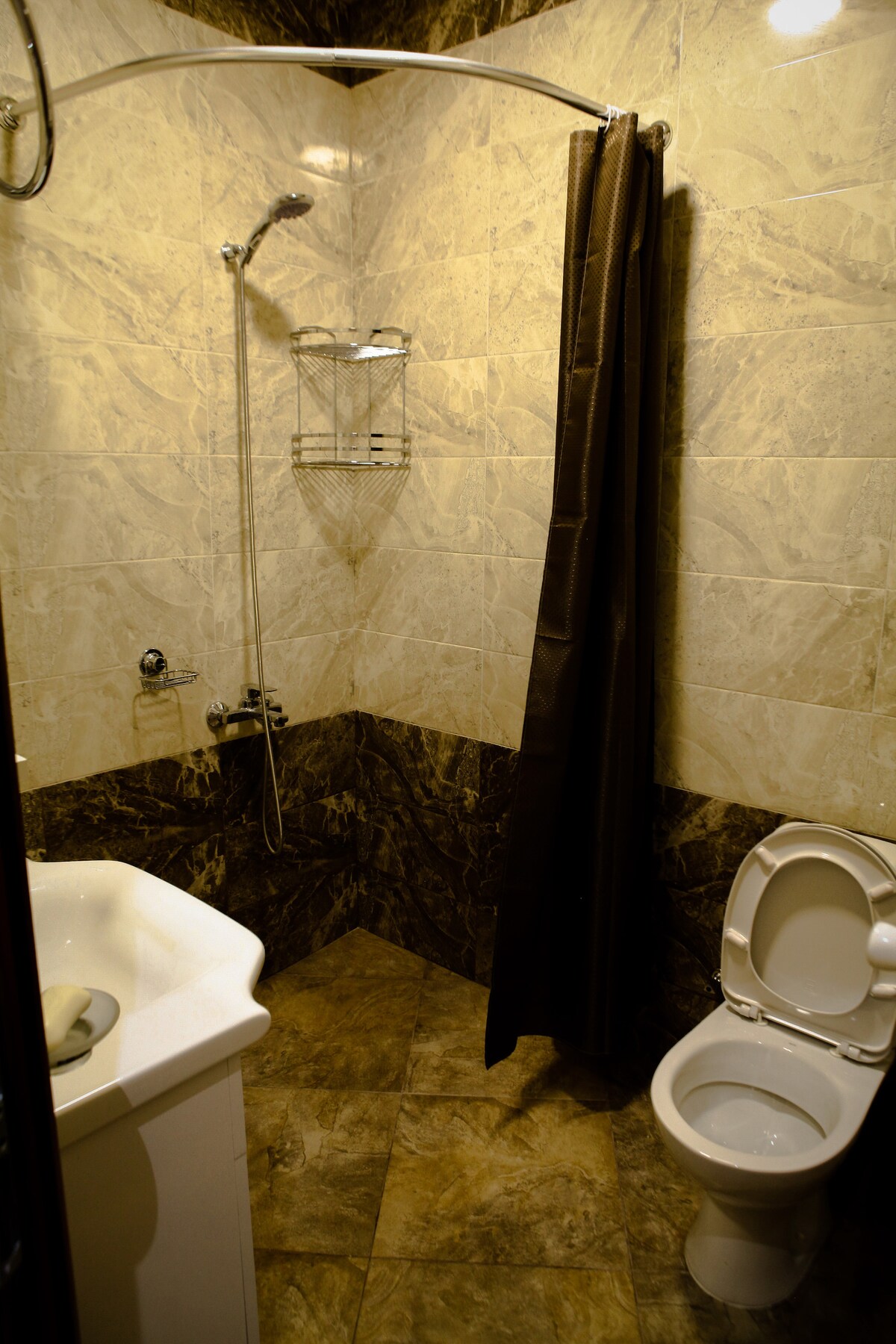 The bathroom features a clean and modern design with beige and brown marbled tiles. A shower area is equipped with a fixed showerhead and a corner shelf. A simple white sink is situated beside a toilet, with a dark curtain providing privacy.