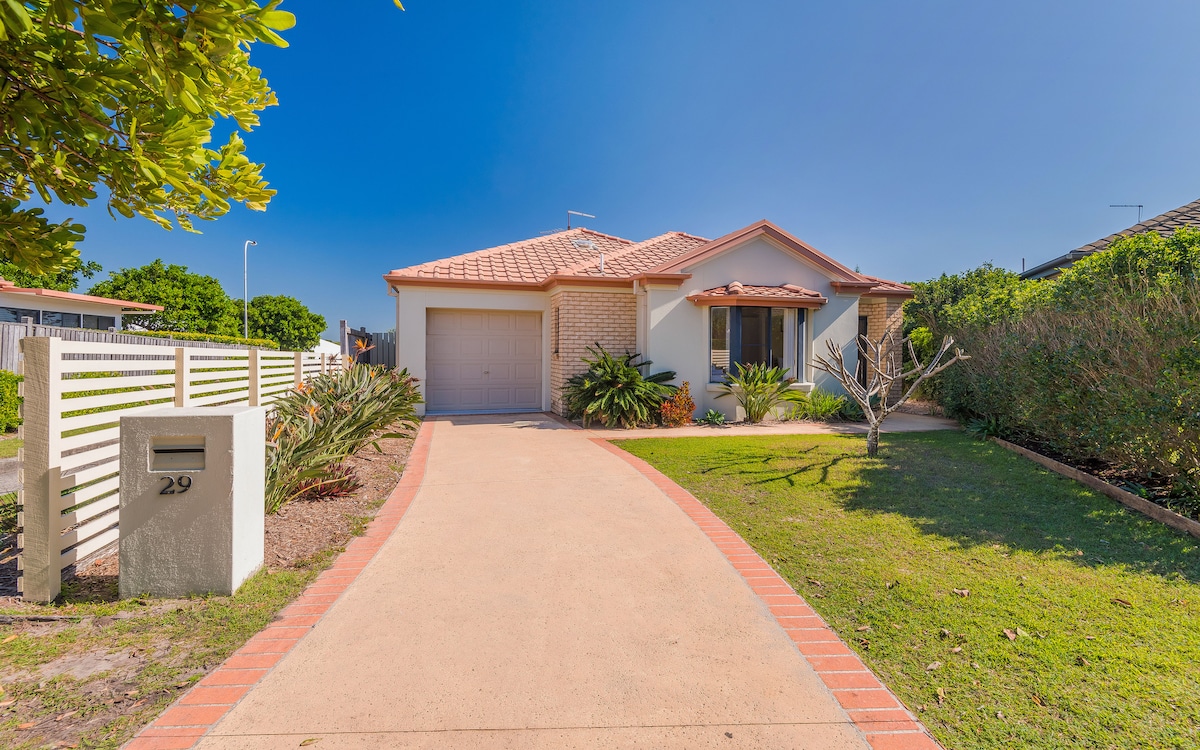 A single-story house is set amidst a well-maintained garden, showcasing a brick-paved driveway leading to a garage. Surrounded by greenery, the front patio includes various tropical plants, and the entrance features a large window allowing natural light to enter the living space.