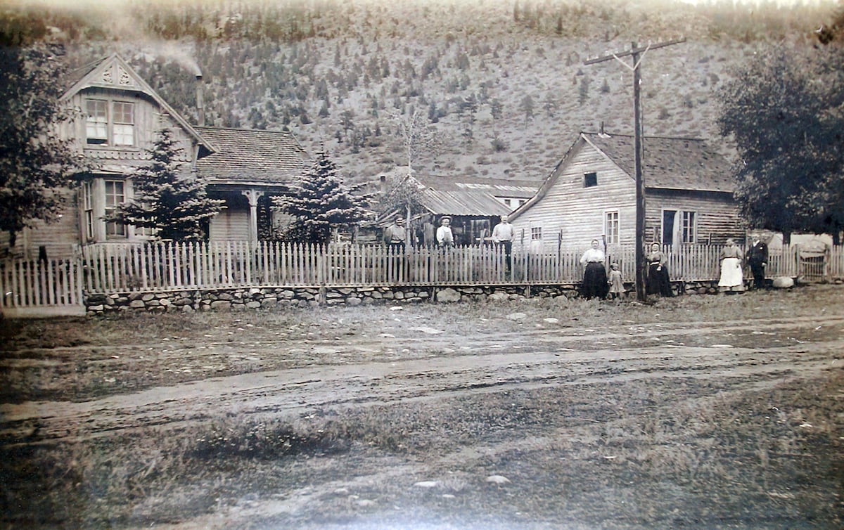A historic photograph captures a Victorian house surrounded by a white picket fence. Several individuals are seen outside, engaging in daily activities. The landscape features a rugged mountain backdrop, with trees and grassy areas enhancing the serene setting.